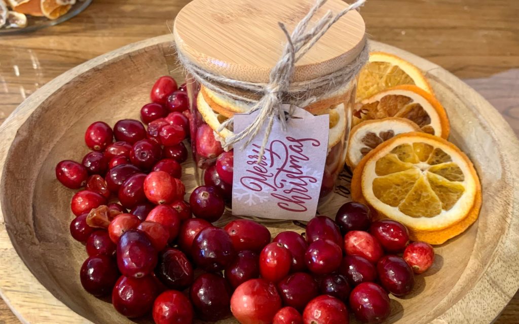 A wooden tray with cranberries, oranges and a mason jar sitting on it. The mason jar has a Merry Christmas tag and is filled with potpourri. 