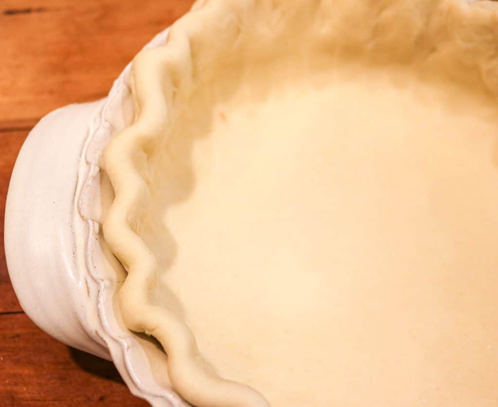 one half of an unbaked pie crust with a fluted edge in a white pie pan on a wooden counter
